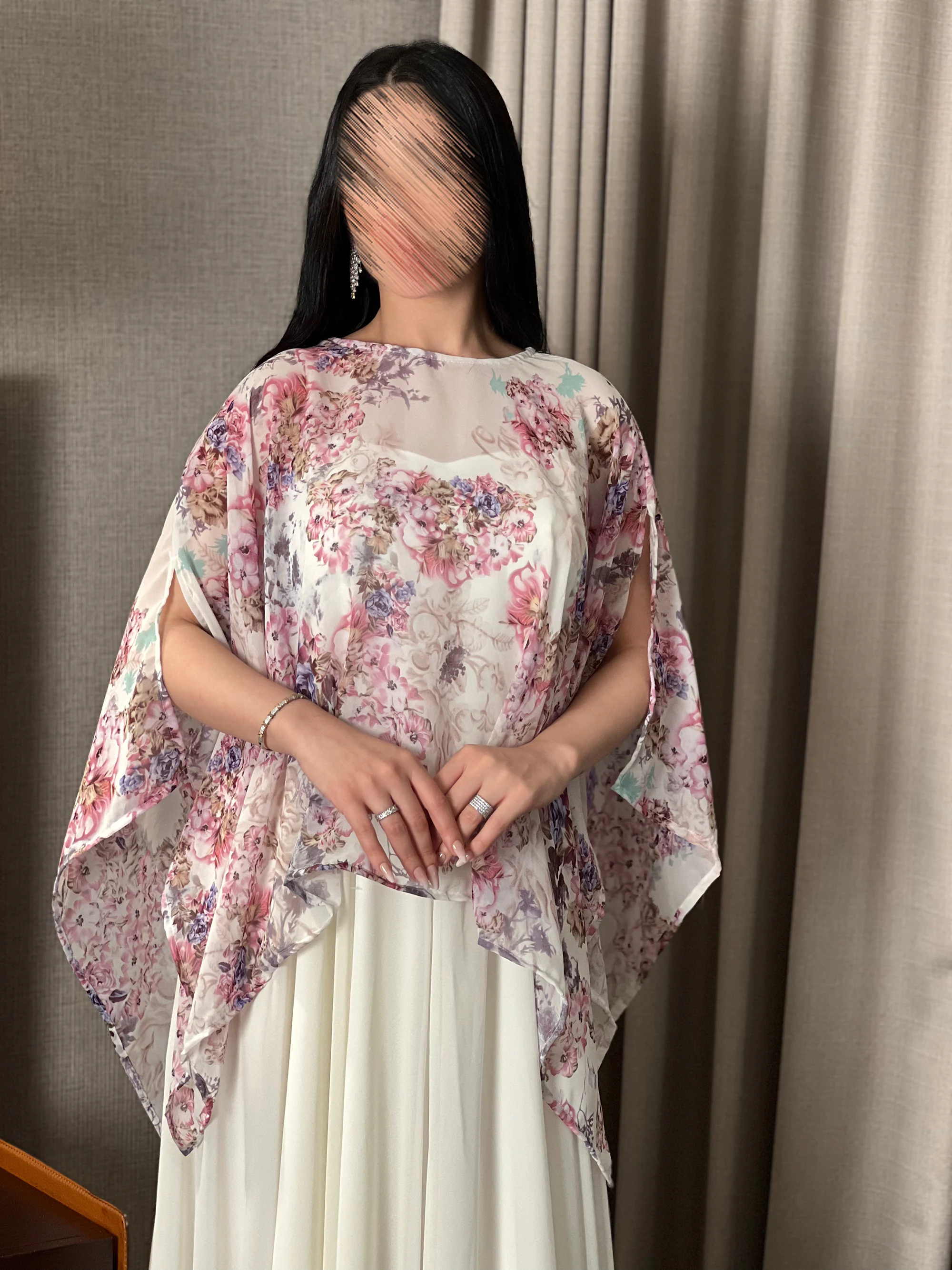 إطلالة أمامية تبرز تصميم الكب (Cape) المورد المنسدل فوق فستان الجورجيت بكسراته المتقنة.