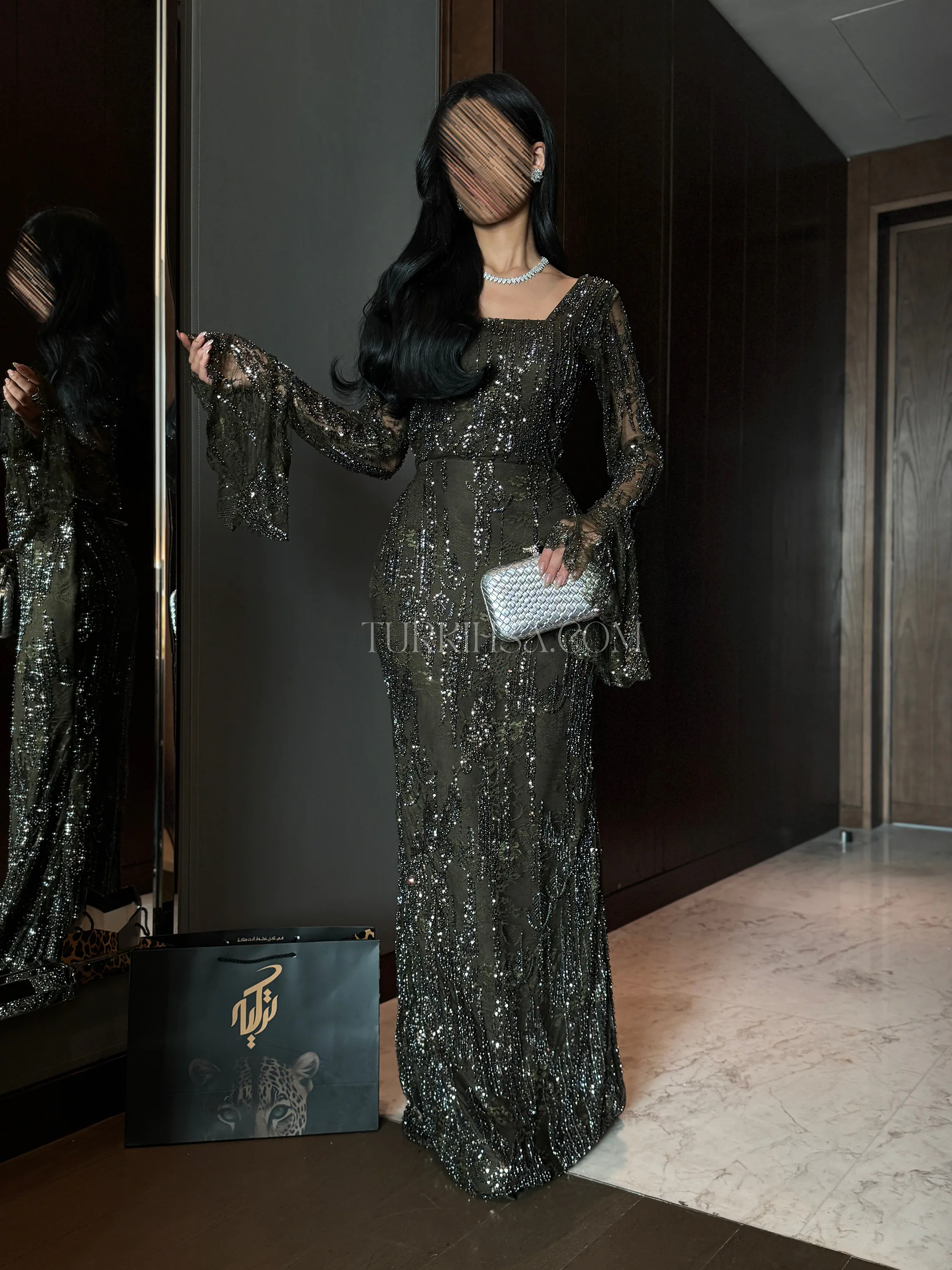إطلالة أمامية كاملة بوضعية الوقوف تبرز فخامة اللون (الزيتي/البرونزي) المطرز، وتوضح قصة الأكمام الطويلة الواسعة من الأسفل (Bell sleeves) التي تضفي لمسة كلاسيكية راقية، مع ظهور حقيبة متجر 