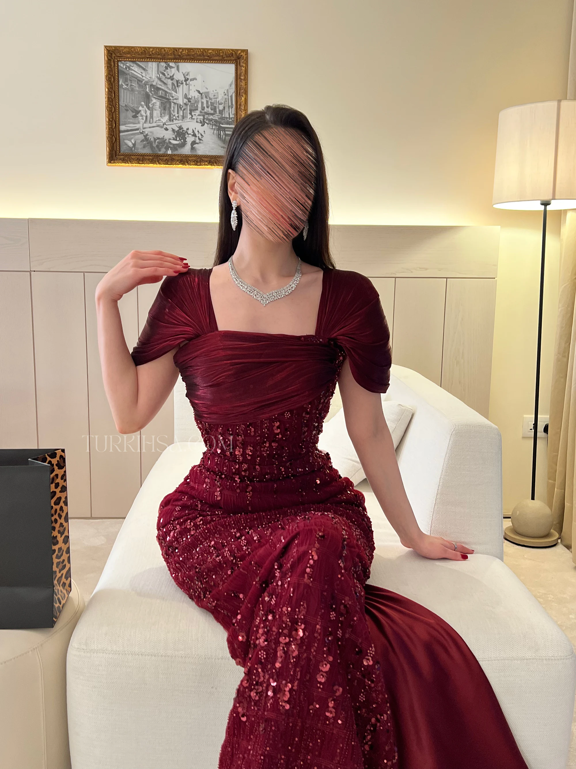 تُظهر انسيابية الفستان من الأسفل وظهور الذيل الساتان الناعم، وتبرز تفاصيل الأكمام والياقة في وضعية مريحة وفخمة بجوار النافذة.