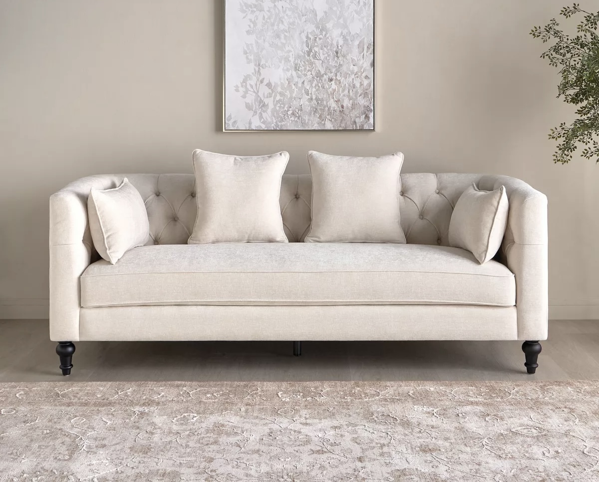 A beige tufted sofa with buttons is decorated with four beige cushions and round black legs